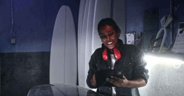 Μαύρη γυναίκα χαμογελά και εργάζεται σε tablet σε surfboard εργαστήριο — Αρχείο Βίντεο