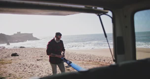 Старший мужчина привязывает поводок к доске для серфинга на пляже — стоковое видео
