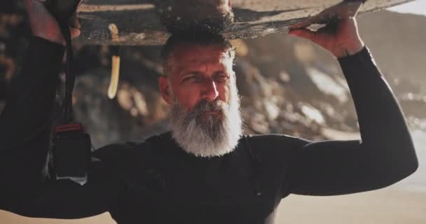 Zbliżenie się starszy przystojny mężczyzna niosąc deska surfingowa na plaży — Wideo stockowe