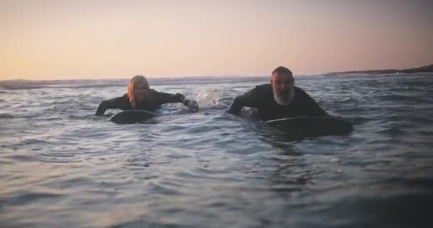 Hombre y mujer mayores surfeando en el agua al atardecer — Vídeo de stock