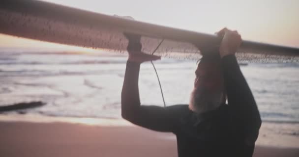 Blisko starszy mężczyzna niosący deskę surfingową na plaży podczas zachodu słońca — Wideo stockowe