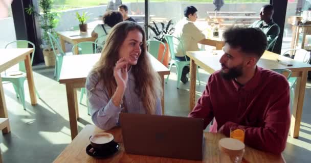 Νεαρή ξανθιά γυναίκα και μεσανατολίτης άντρας που χρησιμοποιεί φορητό υπολογιστή στο καφέ — Αρχείο Βίντεο
