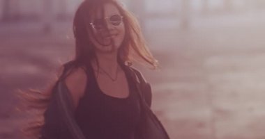 Gün batımında şehirde yürüyen kaykaylı gülümseyen Asyalı kadın.