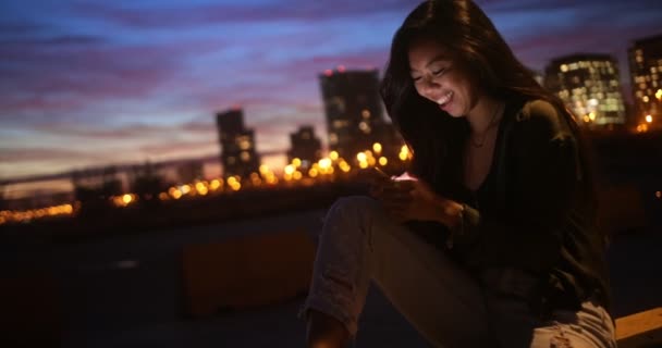 Χαμογελώντας ασιατική γυναίκα γραπτών μηνυμάτων με φώτα της πόλης στο παρασκήνιο — Αρχείο Βίντεο