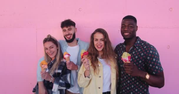 Amici multietnici che tengono gelati colorati contro il muro rosa — Video Stock