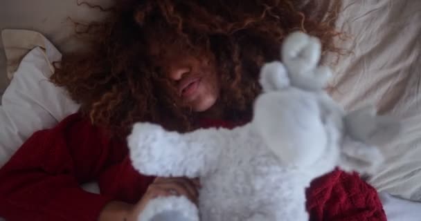 Μαύρη γυναίκα ξαπλωμένη στο κρεβάτι χαμογελώντας παίζοντας με βελούδινο παιχνίδι — Αρχείο Βίντεο