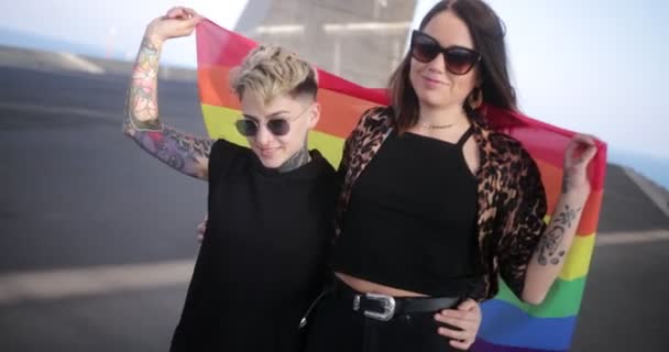 Pareja de lesbianas abrazándose mutuamente sosteniendo la bandera del arco iris del orgullo LGBT — Vídeos de Stock