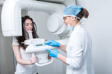 Tıp üniformalı kadın gülümseyerek modern makinede düğmelere basarken klinikteki kadın müşterinin dişlerinin röntgenini çekiyor.