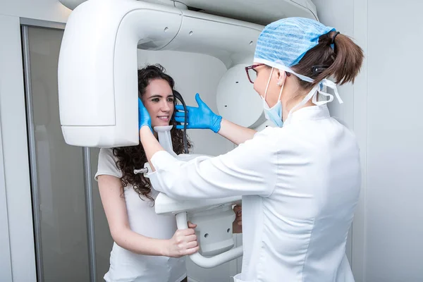 Tıbbi üniformalı kadın doktor, çağdaş diş kliniğinde çalışırken röntgen makinesindeki gülümseyen bayanın başını ayarlıyor.