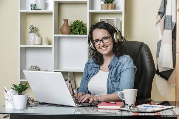Cheerful female freelancer in glasses and wireless headphones wearing casual clothes smiling at camera while sitting in soft leather armchair at table with mug of hot drink and interacting with netbook in contemporary apartment