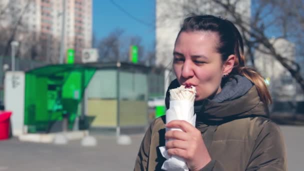Una ragazza per strada mangia un donatore. La ragazza mangia fast food vicino alla strada. Giornata di sole . — Video Stock