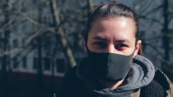 Девушка в чёрной защитной маске смотрит в камеру. На улице, на фоне многоэтажных зданий на заднем плане — стоковое видео