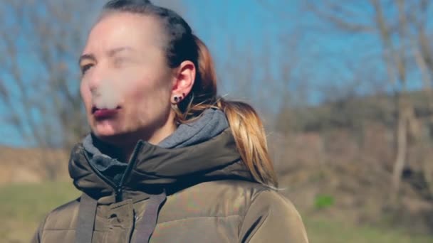 Hermosa joven morena fuma en el parque de otoño y vapeo, al final mirando a la cámara — Vídeos de Stock
