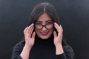 Siyah arka planda siyah ceketi olan ve gözlük takan Latin bir kadın.