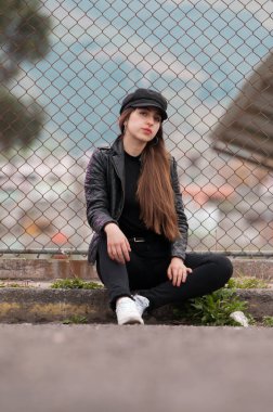 Amerikan portresi, siyah giysiler içinde uzun saçlı ve deri ceketli genç bir kadın oturuyor ve raflara yaslanıyor.