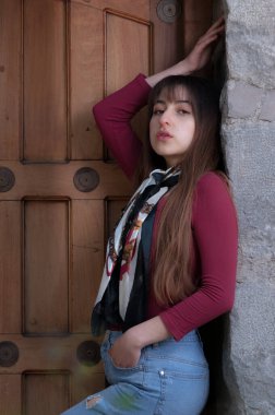 Bir eli belinde, diğer eli de kapı çerçevesinde ahşap kapıya dayanmış uzun saçlı genç bir kadının Amerikan portresi.