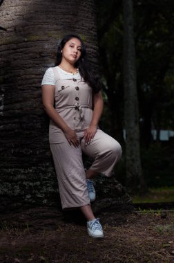 Genç siyah saçlı latin, siyah arka planı olan bir ağaca yaslanıyor.