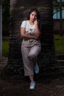 Genç siyah saçlı latin, siyah arka planı olan bir ağaca yaslanıyor.