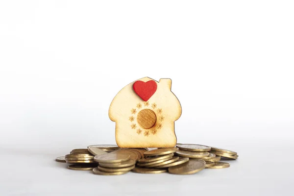 Small wooden house shape with red heart stands on hill of coins, white background. Savings money on utility bills, rent, buying property, real estate investment, mortgage loan concept. Copy space.