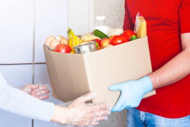 Kırmızı üniformalı ve tıbbi eldivenli kurye kapıya yakın koruyucu eldivenli kadınlara yiyecek kutusu veriyor. Kilitleme, çevrimiçi alışveriş veya bağış konsepti sırasında güvenli gıda teslimatı.