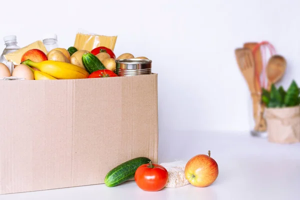 Various grocery items in cardboard box on white table in kitchen. Food box with fresh vegetables, fruits, cereals, pasta, milk, eggs and canned goods. Food delivery or donation concept.