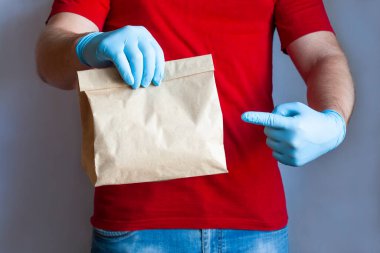 Kırmızı üniformalı ve tıbbi eldivenli kurye paket servis için kağıt yiyecekler tutuyor ve gri arka planda işaret ediyor. Koronavirüs karantinası sırasında güvenli teslimat hizmeti.