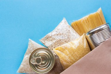 Üst görünümde çeşitli market ürünleri. Makarna, mısır gevreği ve konserve yiyecekler mavi arka planda kese kağıdında. Yiyecek teslimatı, bağış veya hisse senedi konsepti. Boşluğu kopyala.