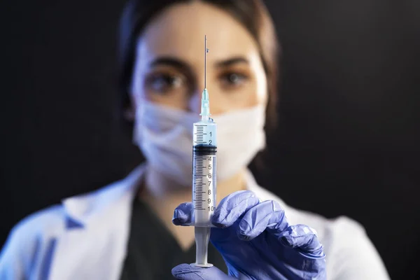 Caucasian white Doctor or nurse woman with mask and blue examination gloves holds injection syringe and vaccine on a black background.