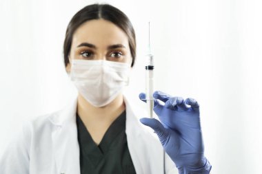 Maskeli ve mavi eldivenli beyaz doktor ya da hemşire kadın beyaz arka planda şırınga ve aşı tutuyor..