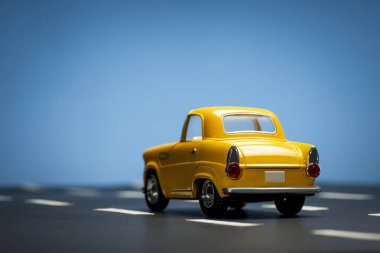 Mavi arka planda ve asfalt yolda sarı bir oyuncak arabanın arka görüntüsü.