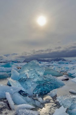 Işıl ışıl mavi buzdağları bulutlu bir günde Jokulsarlon Okyanus Gölünde yüzer. Arka planda dağlar ve yukarıda güneş bulunur.