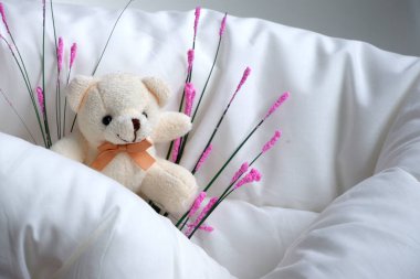 Ayı, yastık ve battaniyeden yapılmış yumuşak bir yatakta oturuyor ve yanında çiçekler ile güneşleniyor. Yumuşak oyuncak. Ayı yumuşak bej