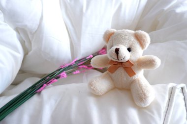 Ayı, yastık ve battaniyeden yapılmış yumuşak bir yatakta oturuyor ve yanında çiçekler ile güneşleniyor. Yumuşak oyuncak. Ayı yumuşak bej