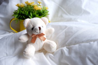 Yumuşak bej bir ayı, pamuk beyazı çarşafların üzerinde oturuyor, çiçeklerin arka planında. Çocuk oyuncağı..