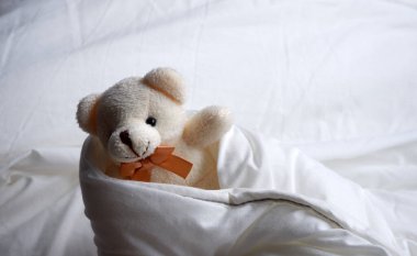 Beyaz bir battaniyeye sarılı bej bir ayı elini sallıyor. Çocuk oyuncağı.