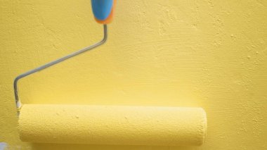 Mavi eldiven yavaşça beyaz duvarı silindirle sarıya boyuyor. Yakın çekim görünümü