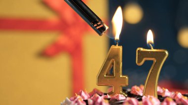 Birthday cake number 47 golden candles burning by lighter, background gift yellow box tied up with red ribbon. Close-up