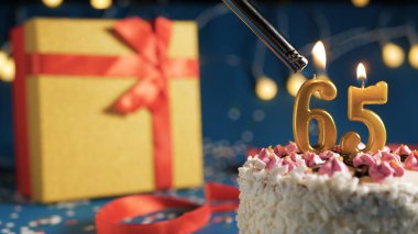 White birthday cake number 65 golden candles burning by lighter, blue background with lights and gift yellow box tied up with red ribbon. Close-up