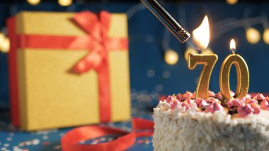 White birthday cake number 70 golden candles burning by lighter, blue background with lights and gift yellow box tied up with red ribbon. Close-up