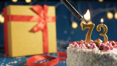 White birthday cake number 73 golden candles burning by lighter, blue background with lights and gift yellow box tied up with red ribbon. Close-up