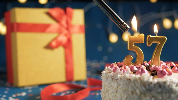 Pastel de cumpleaños blanco número 57 velas doradas encendidas por un fondo azul más claro con luces y caja amarilla de regalo atada con cinta roja. Primer plano —  Fotos de Stock