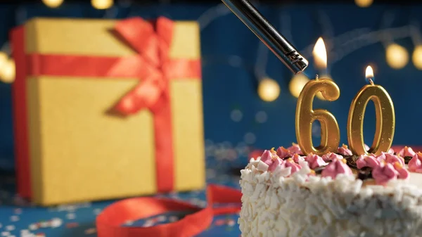 Gâteau d'anniversaire blanc numéro 60 bougies dorées brûlant par briquet, fond bleu avec des lumières et cadeau boîte jaune attaché avec ruban rouge. Gros plan — Photo