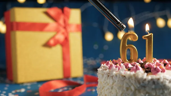 Pastel de cumpleaños blanco número 61 velas doradas encendidas por un fondo azul más claro con luces y caja amarilla de regalo atada con cinta roja. Primer plano —  Fotos de Stock
