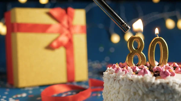 White birthday cake number 80 golden candles burning by lighter, blue background with lights and gift yellow box tied up with red ribbon. Close-up — ストック写真