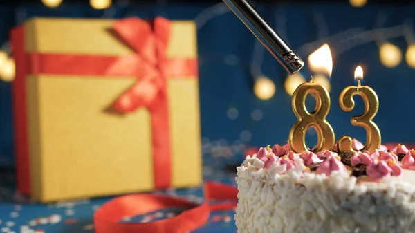 Bolo de aniversário branco número 83 velas douradas queimando por mais leve, fundo azul com luzes e caixa de presente amarelo amarrado com fita vermelha. Close-up — Fotografia de Stock