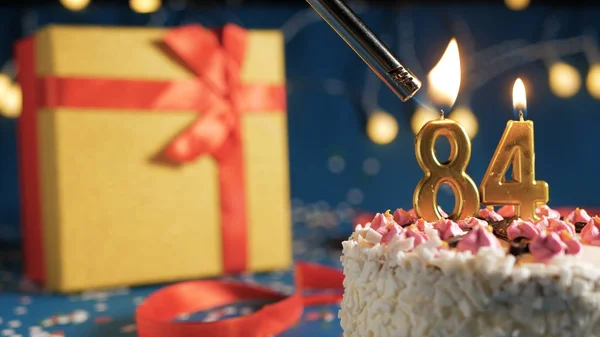 Torta de cumpleaños blanca número 84 velas doradas encendidas por un fondo más claro y azul con luces y caja amarilla de regalo atada con cinta roja. Primer plano —  Fotos de Stock