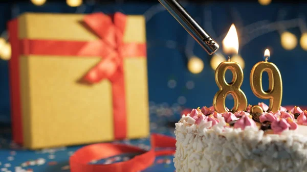 White birthday cake number 89 golden candles burning by lighter, blue background with lights and gift yellow box tied up with red ribbon. Close-up — Stock Photo, Image