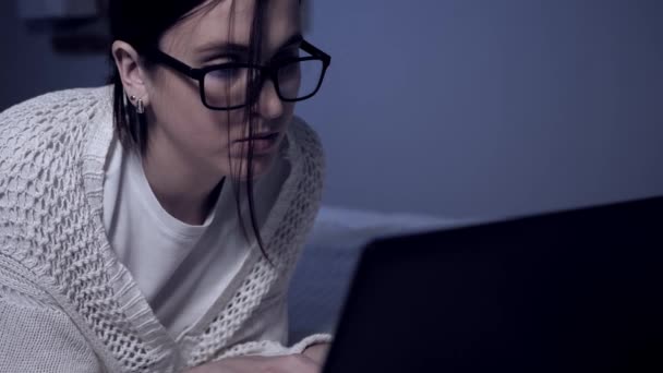 La fille travaille sur un ordinateur portable la nuit. Attrayant jeune étudiante ou pirate ou pigiste avec des lunettes est couché sur le lit sur le ventre et dactylographier quelque chose sur le clavier de l'ordinateur portable la nuit seule dans l'appartement — Video