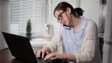İş yerinde serbest çalışan bir kız. Gözlüklü çekici bir kadın rahatsız edici bir pozisyonda telefonla konuşuyor ve aynı zamanda evde mutfaktayken iki eliyle dizüstü bilgisayarda klavye çalıyor. Orta boy.