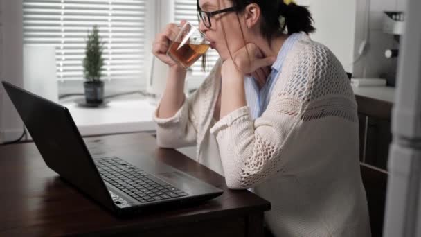 Menina caucasiana atraente em óculos e camisola de malha está na cozinha no apartamento sentado à mesa bebendo chá e olhando para algo na tela do laptop. Mulher freelancer no conceito de trabalho — Vídeo de Stock